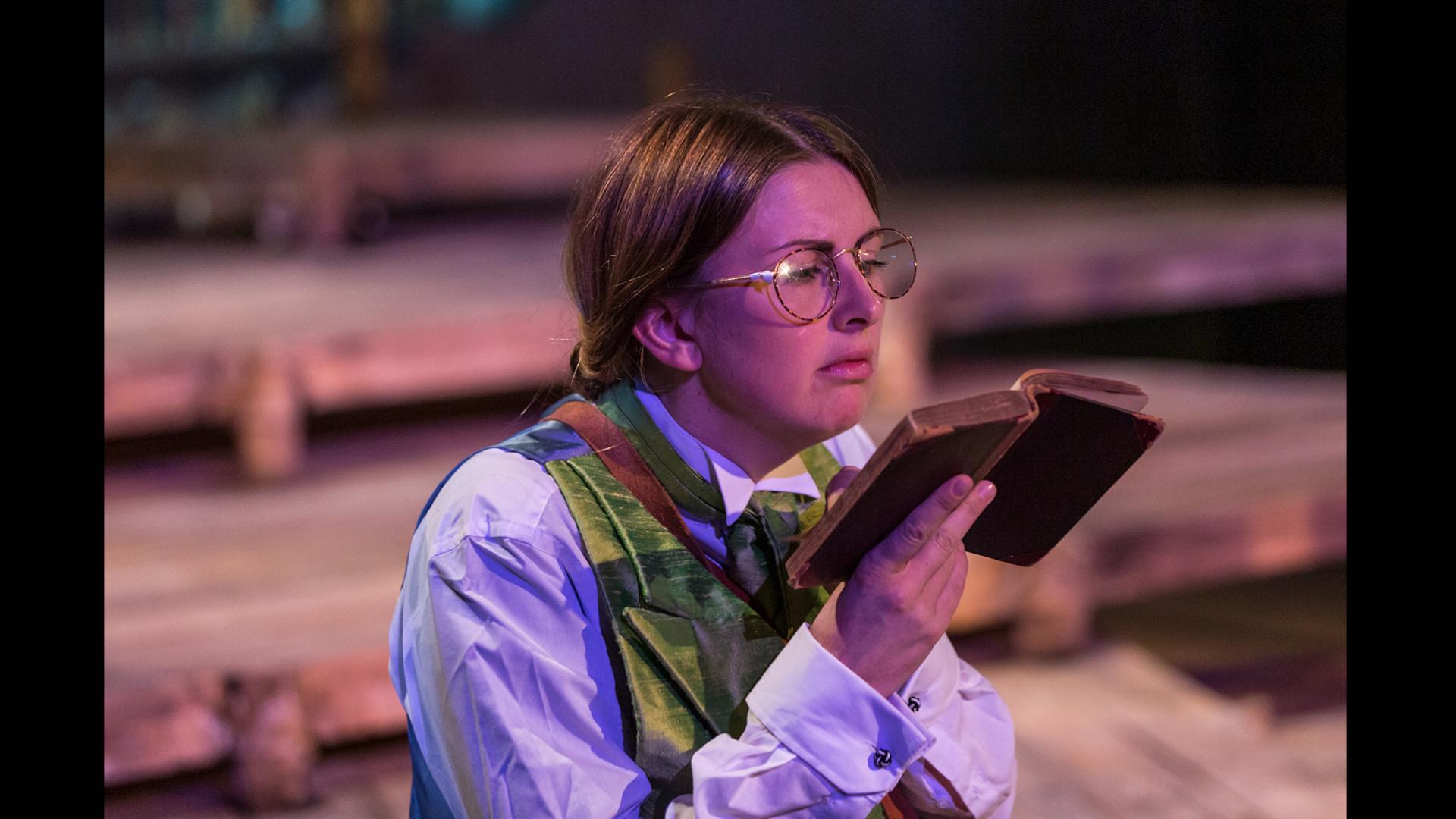 Actor playing Bates, dressed in a green waistcoat, white shirt, wearing wire rimmed glasses and reading an old book.