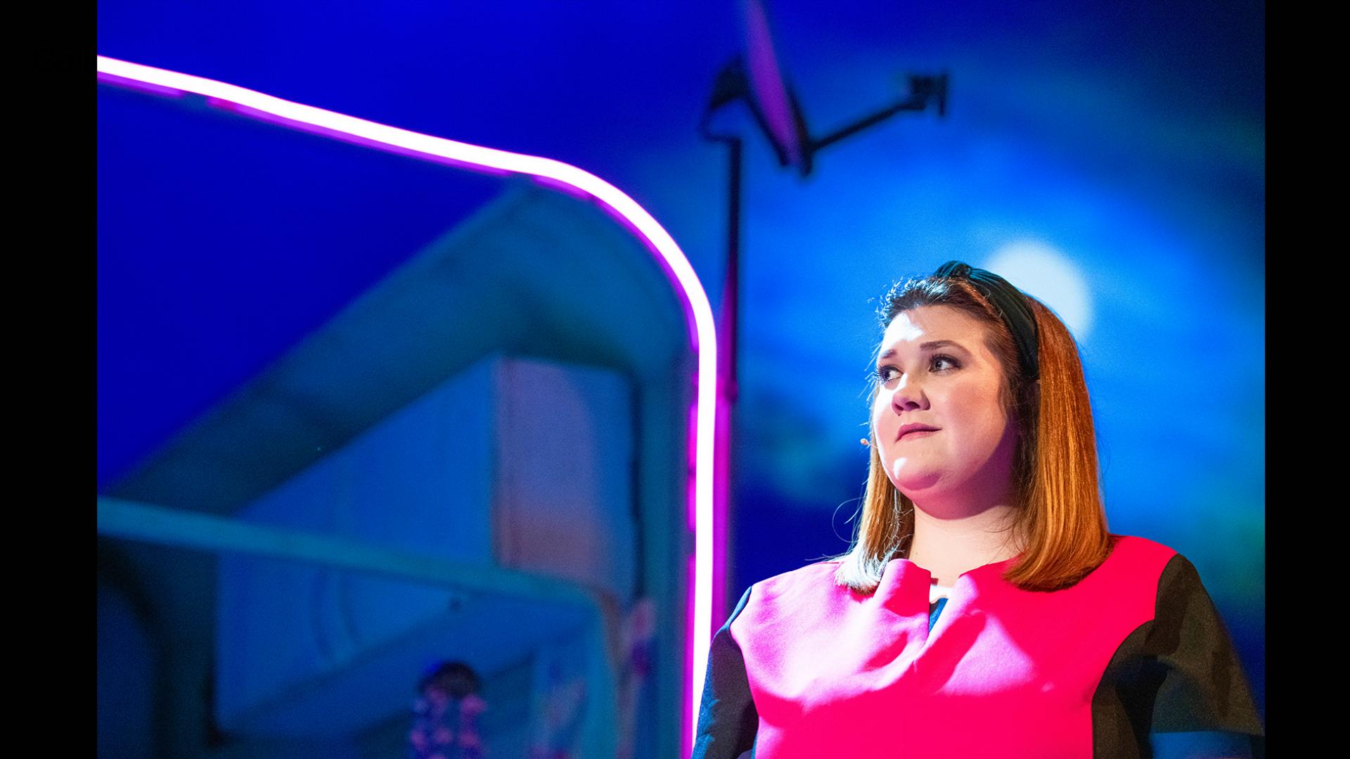 A woman standing in though, wearing a pink tabard with a pink neon light behind