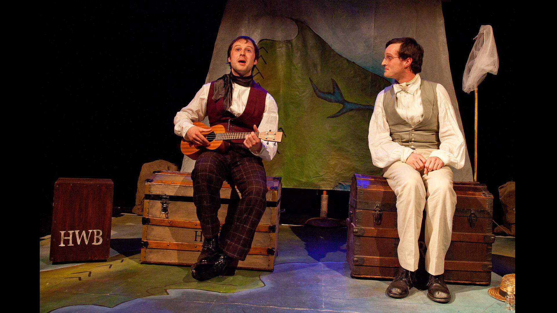 Two actors dressed as Victoria explorers Wallace and Bates, sat on travelling trunks. One is playing a small guitar.