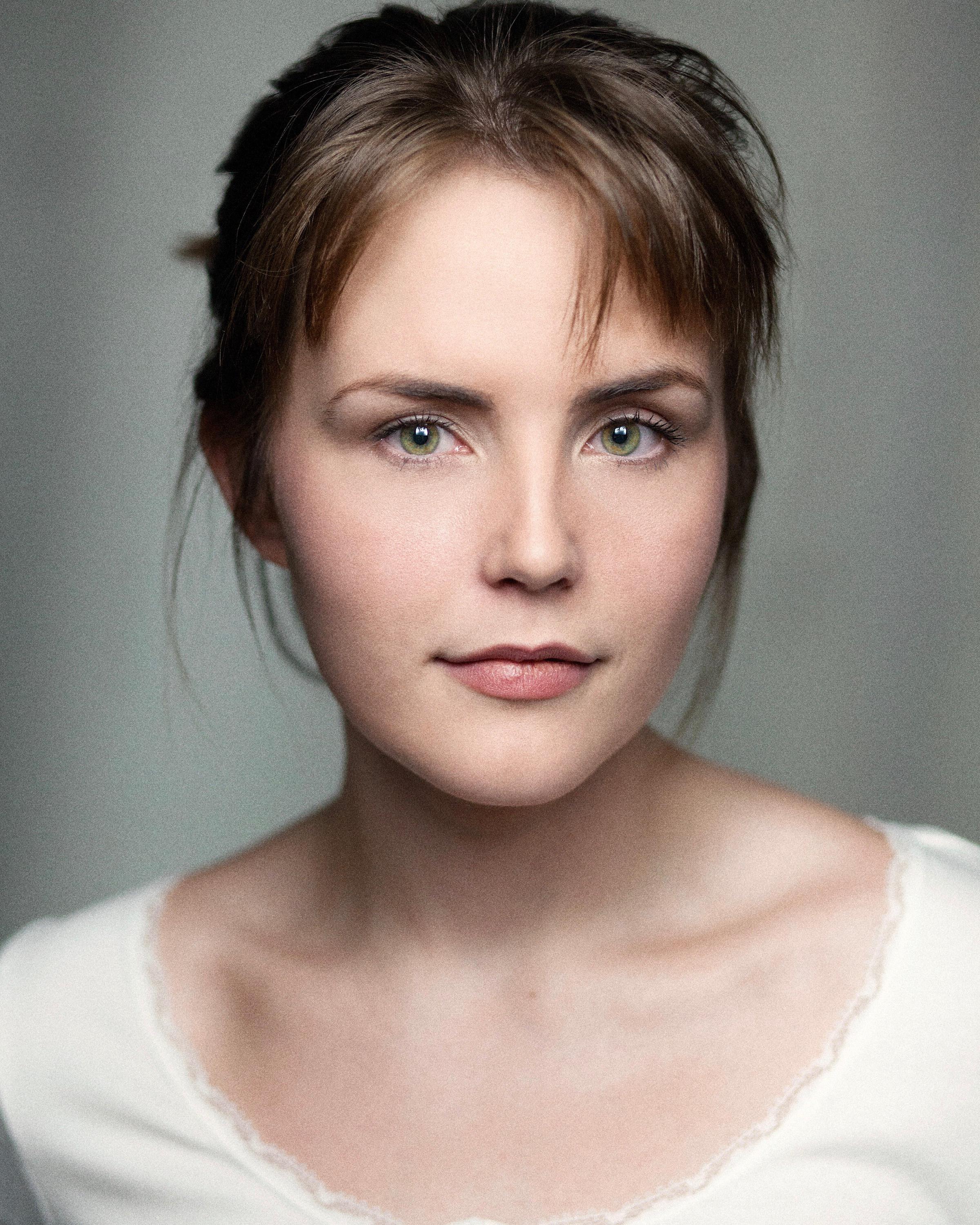Woman with brown hair tied back with a fringe and green eyes wearing a white top