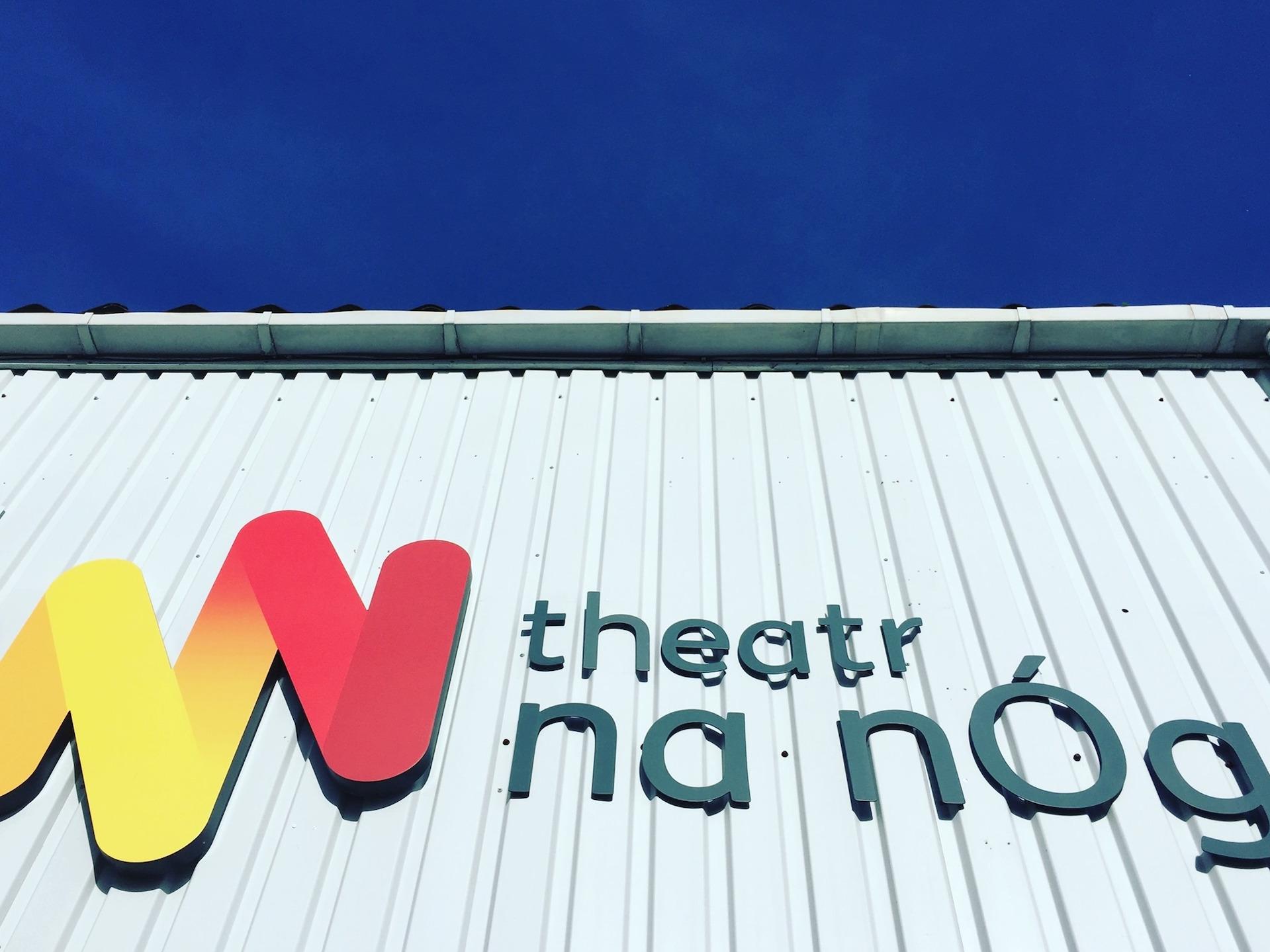 A grey building with the Theatr na nÓg logo and blue sky