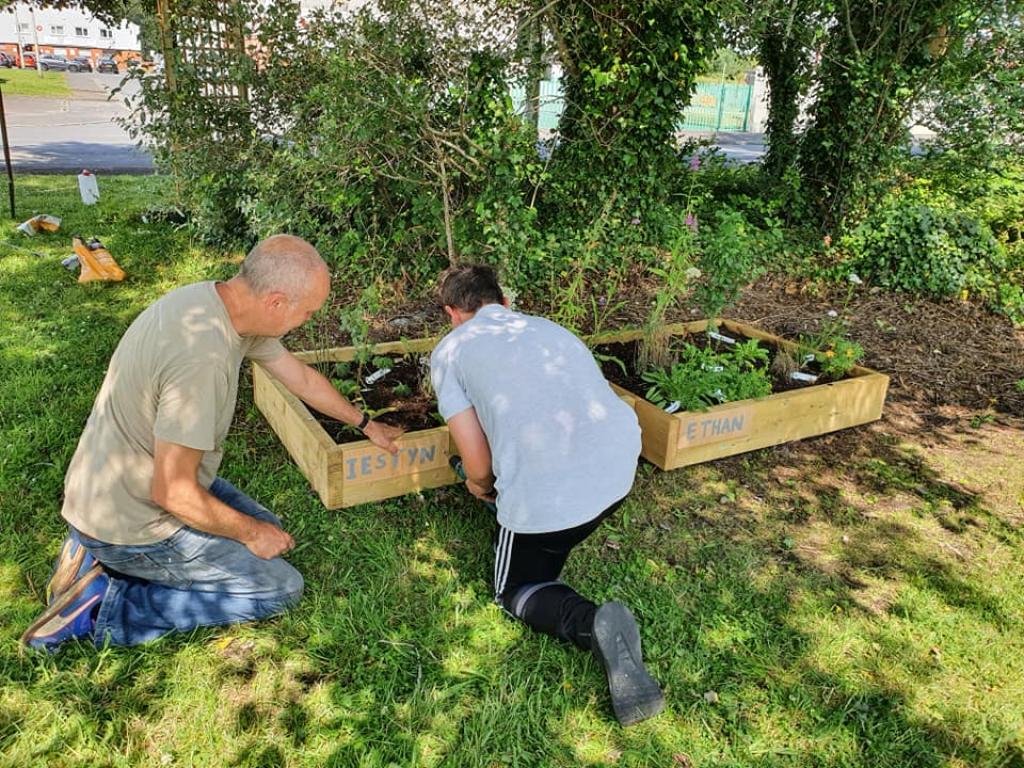 Man in green top and blue jeans kneeling down to help boy in grey top and black trousers to secure a name plaque on to a flower bed in a garden