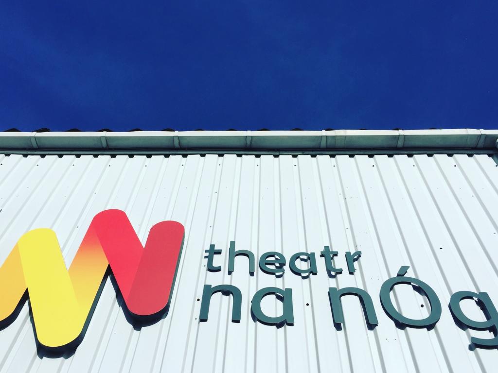 Awyr glas ac adeilad llwyd gyda logo Theatr na nÓg 