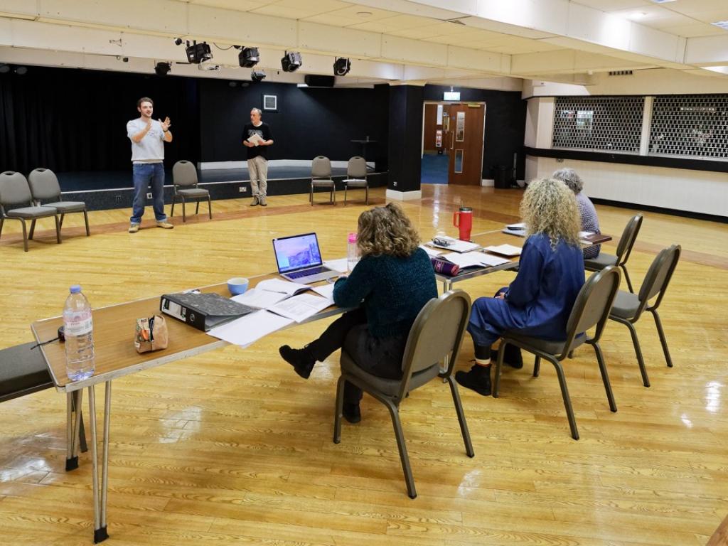 Rehearsal room with two actors and stage managers sat behind a desk