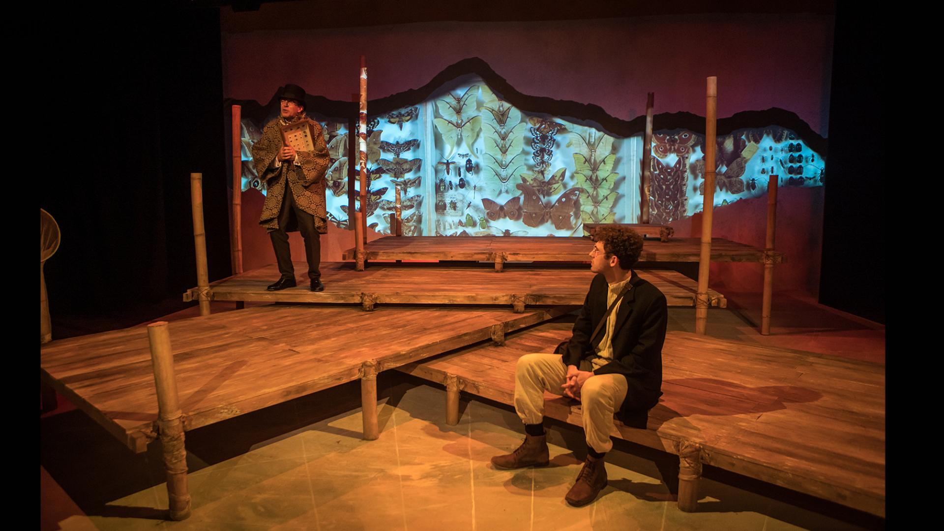 A multi-level stage made of wooden decks. One man sits at the front, another stands at the back. There is a backdrop with images of butterflies.