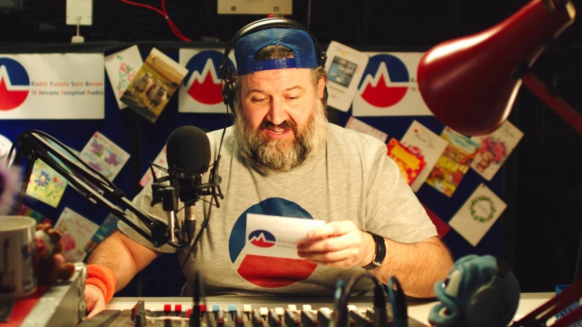 A man in a radio studio sat a mixing desk reading from a card