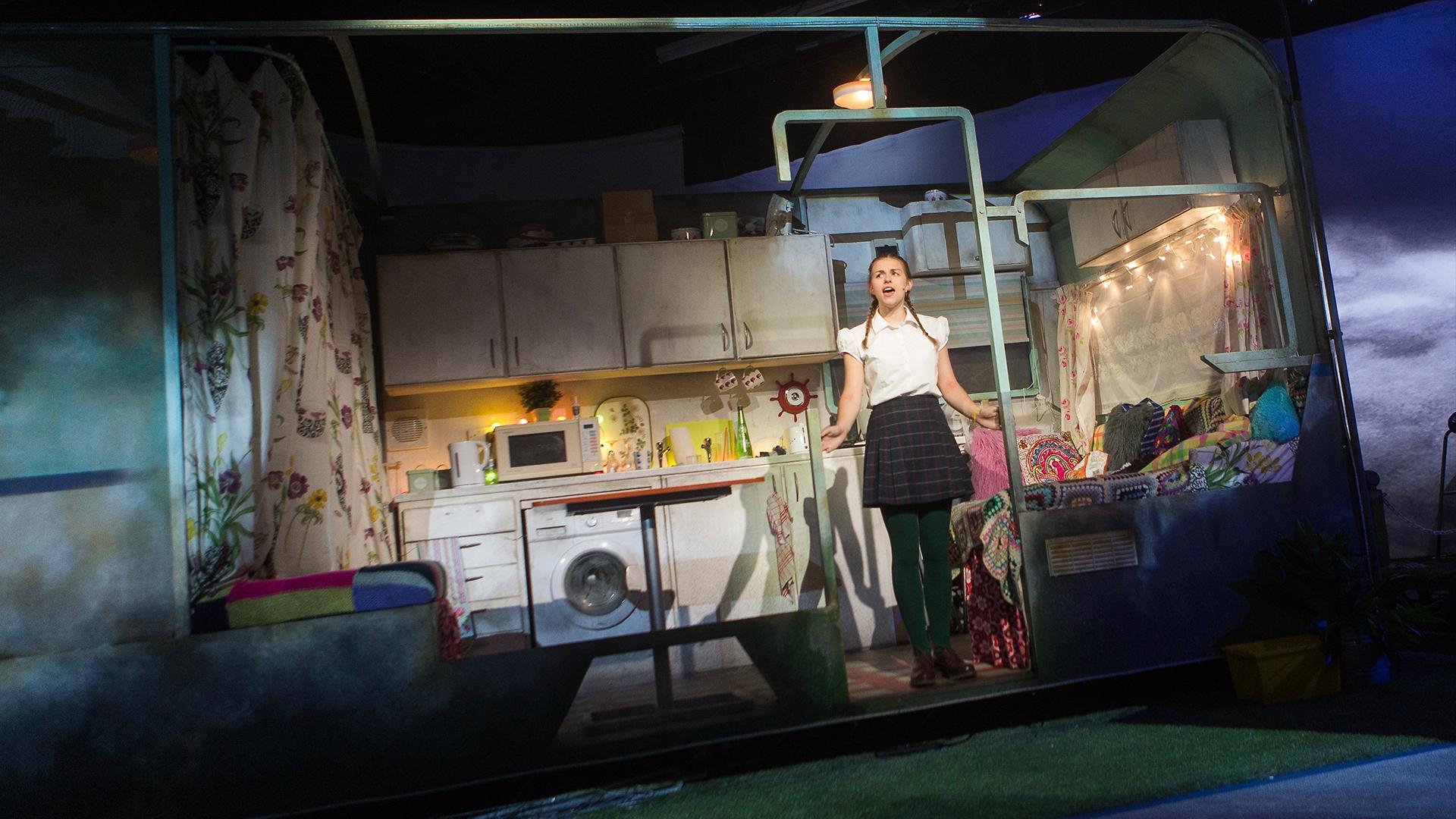 An actor dressed as a school girl standing on the set in the doorway of a caravan with the front removed