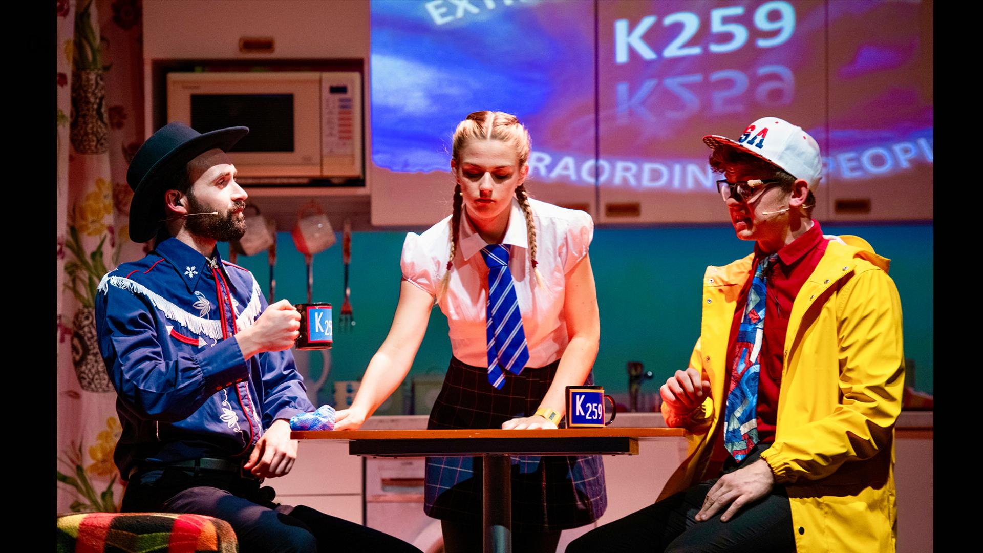 Three people at a table, one dressed as a cowboy, one in a yellow rain mac and a school girl cleaning the table between them.