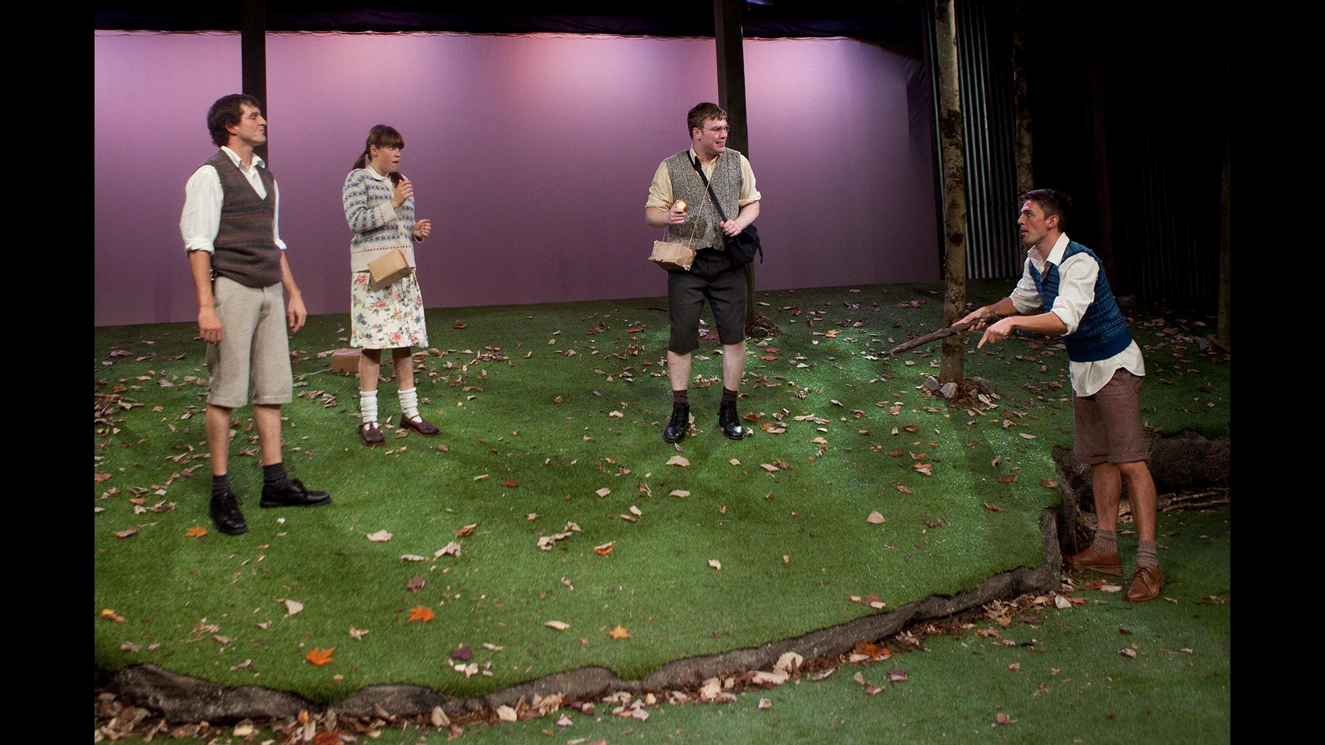 Four actors dressed as children in 1945, standing on a stage covered in grass.