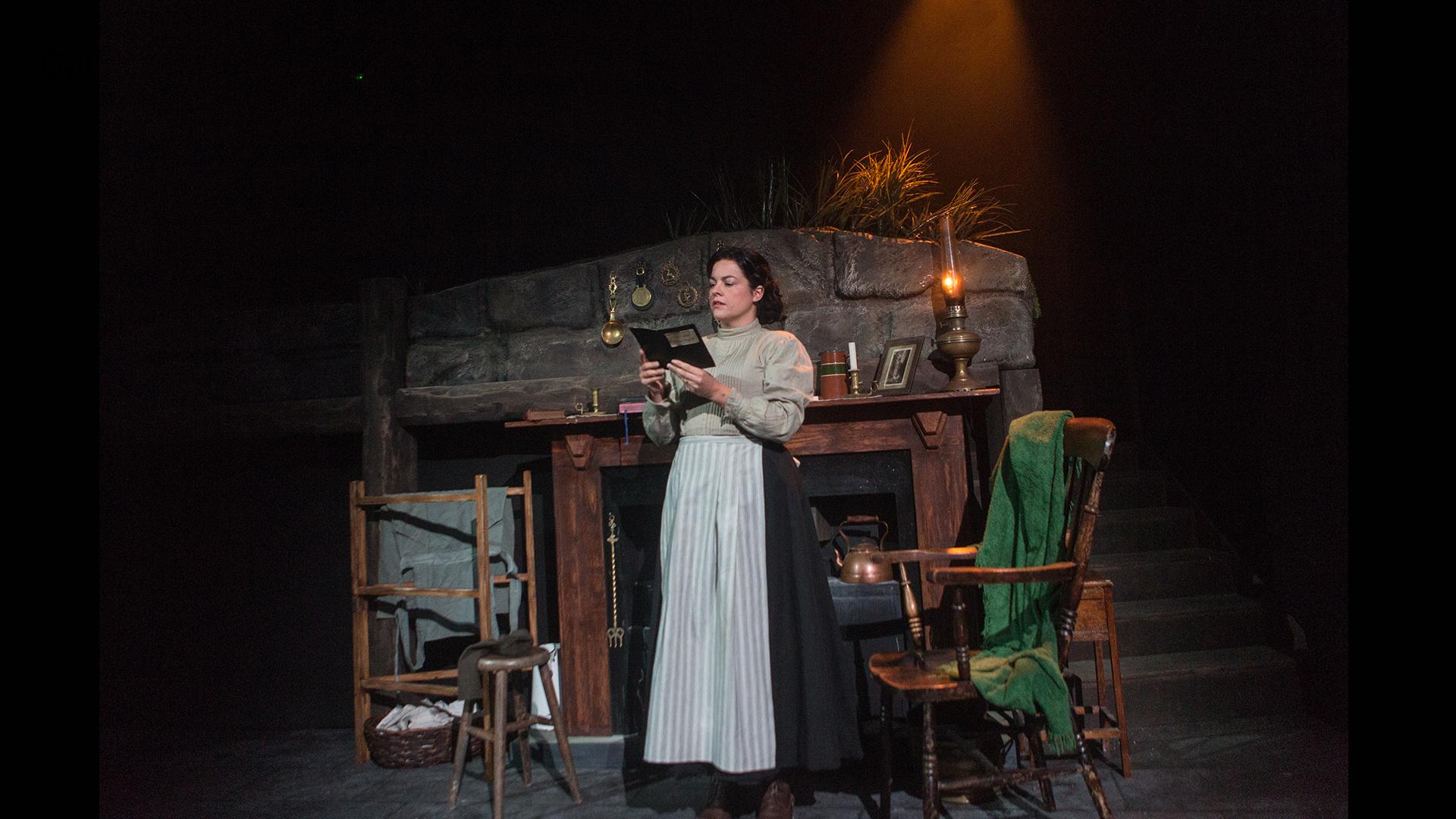 A young woman standing holding a book in front of a fireplace, dressed in a long black skirt with white apron