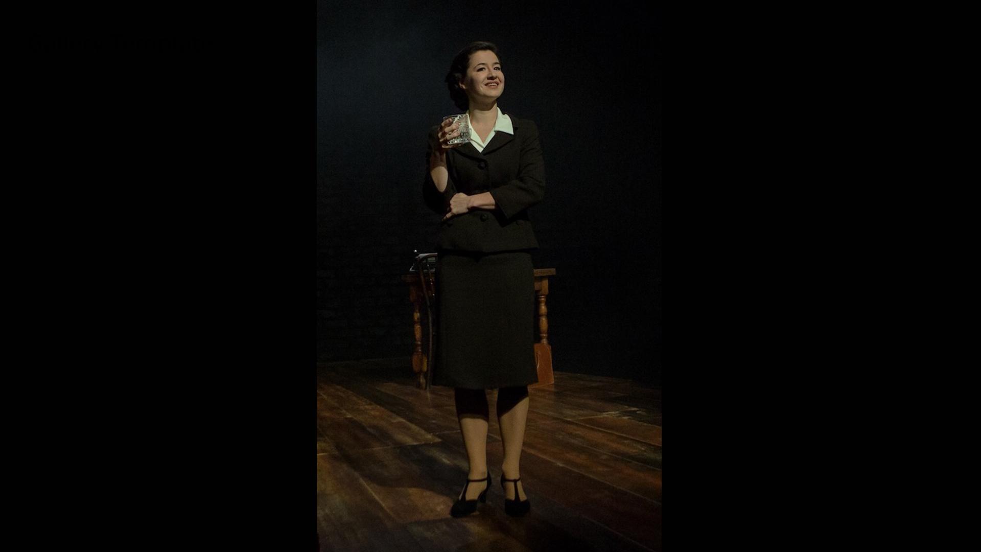 An actor depicting Jennie Lee standing holding a crystal drinking glass