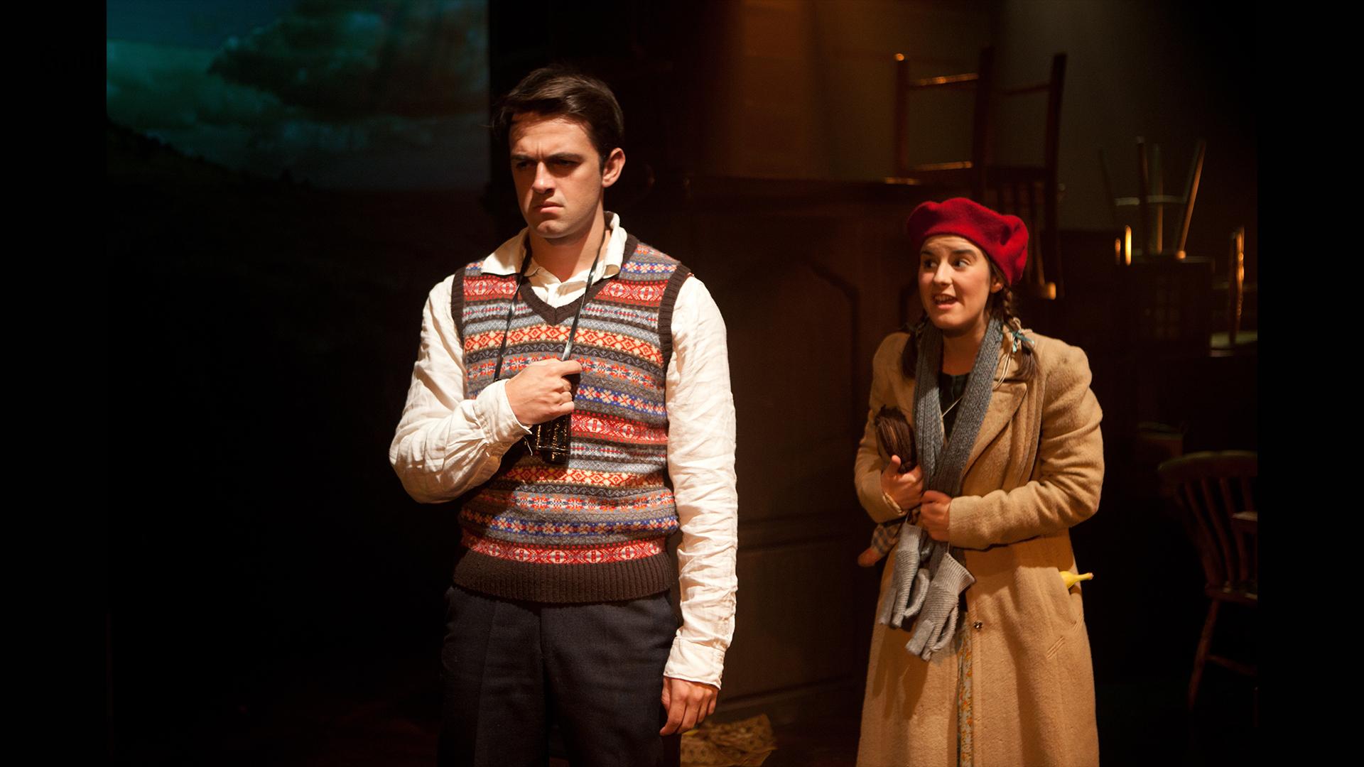 A young man wearing a white shirt and coloured tank top with a girl in a red hat and beige overcoat.