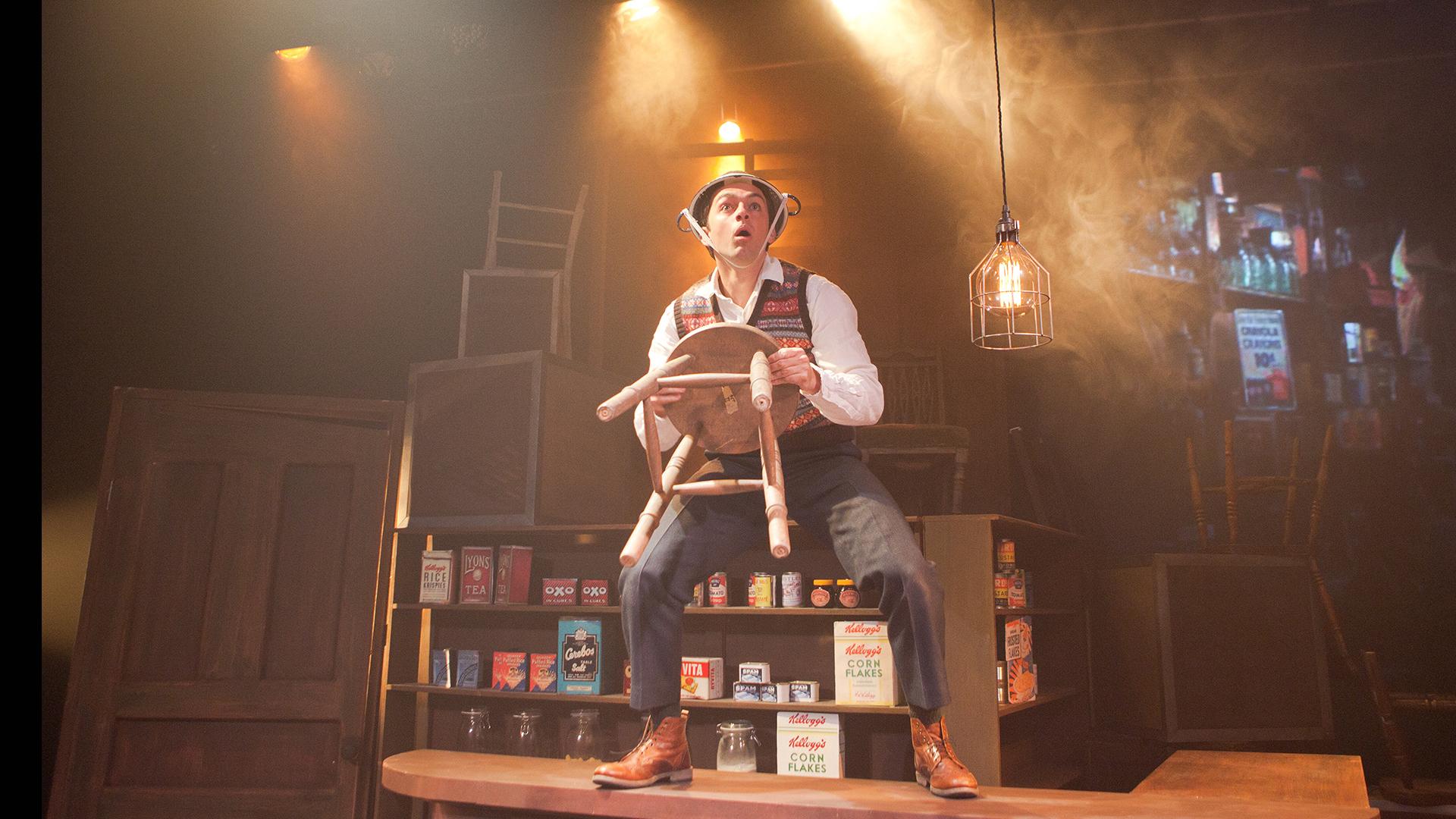 A young man standing on top of an old fashioned shop counter, holding a stool and wearing a colander on his head. 