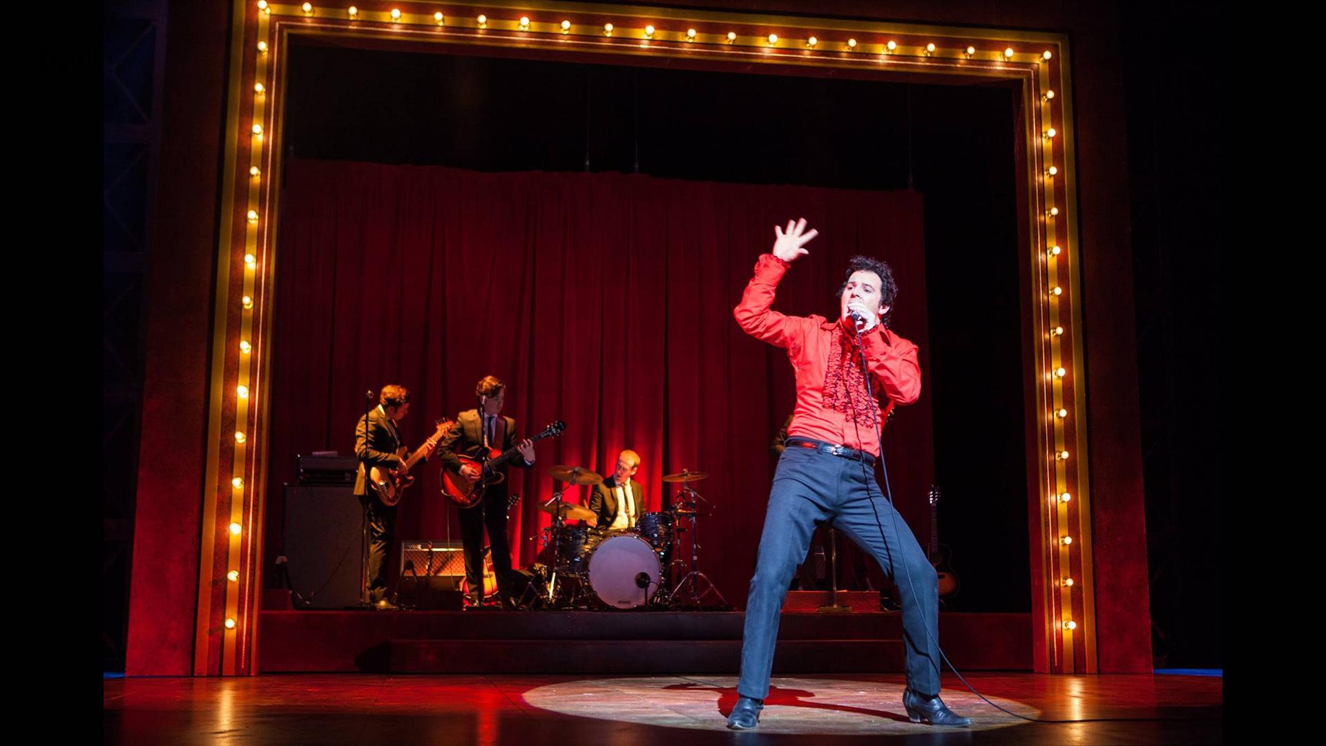 Actor dressed as Tom Jones performing on stage with lights all around and band behind