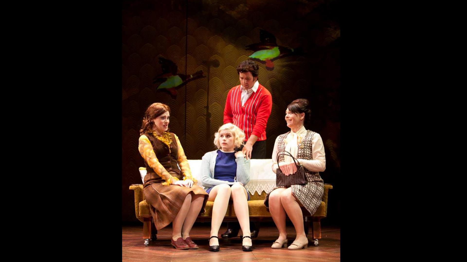 Three women sat on a sofa with a man standing behind. All are dressed in clothes from the 1960s.