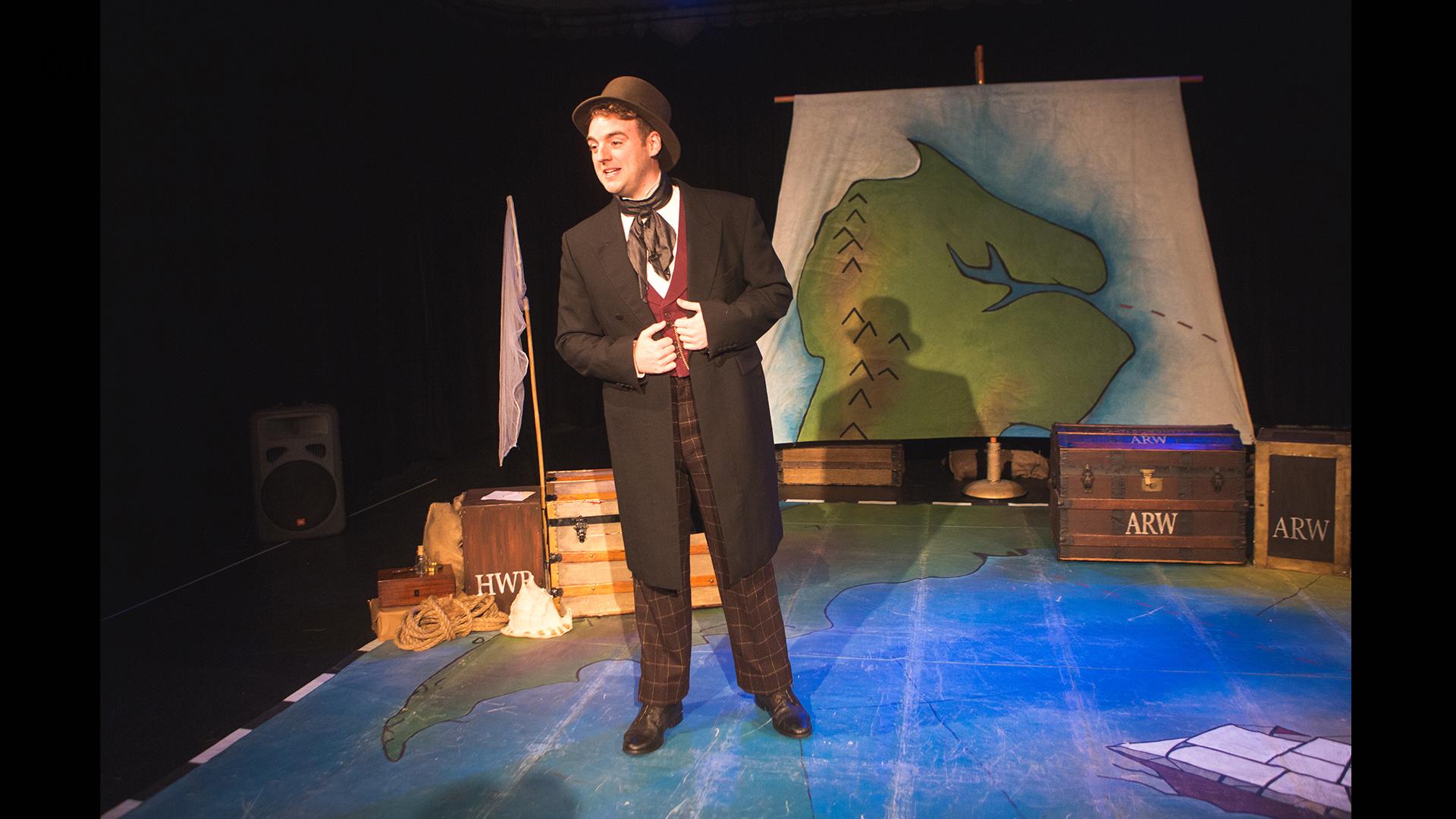 A man dressed in a suit and top hat standing on a stage with wooden travel trunks 