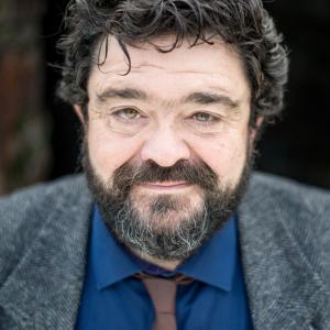 Man with black curly hair and facial hair wearing a blue shirt with a brown tie and a grey jacket