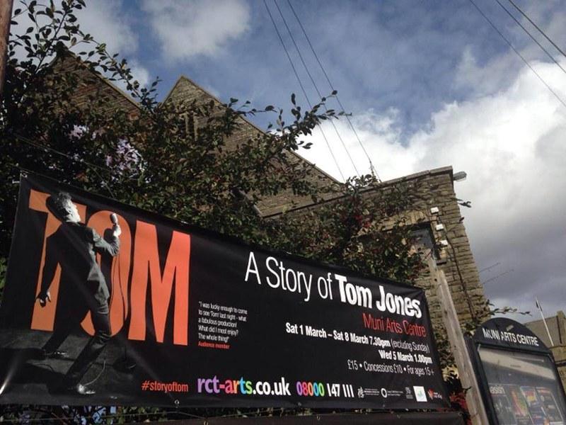 Photograph of a poster for 'A Story of Tom Jones' outside Muni Arts Centre - the poster consists of a red graphic with the word 'TOM' and an actor in front of it from behind singing into a microphone dressed as Tom Jones.