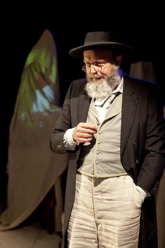 Image of Alfred Russel Wallace in waistcoat and white trousers, with a black tailcoat, hat and round glasses looking to the side. Blurred colours are visible in background.