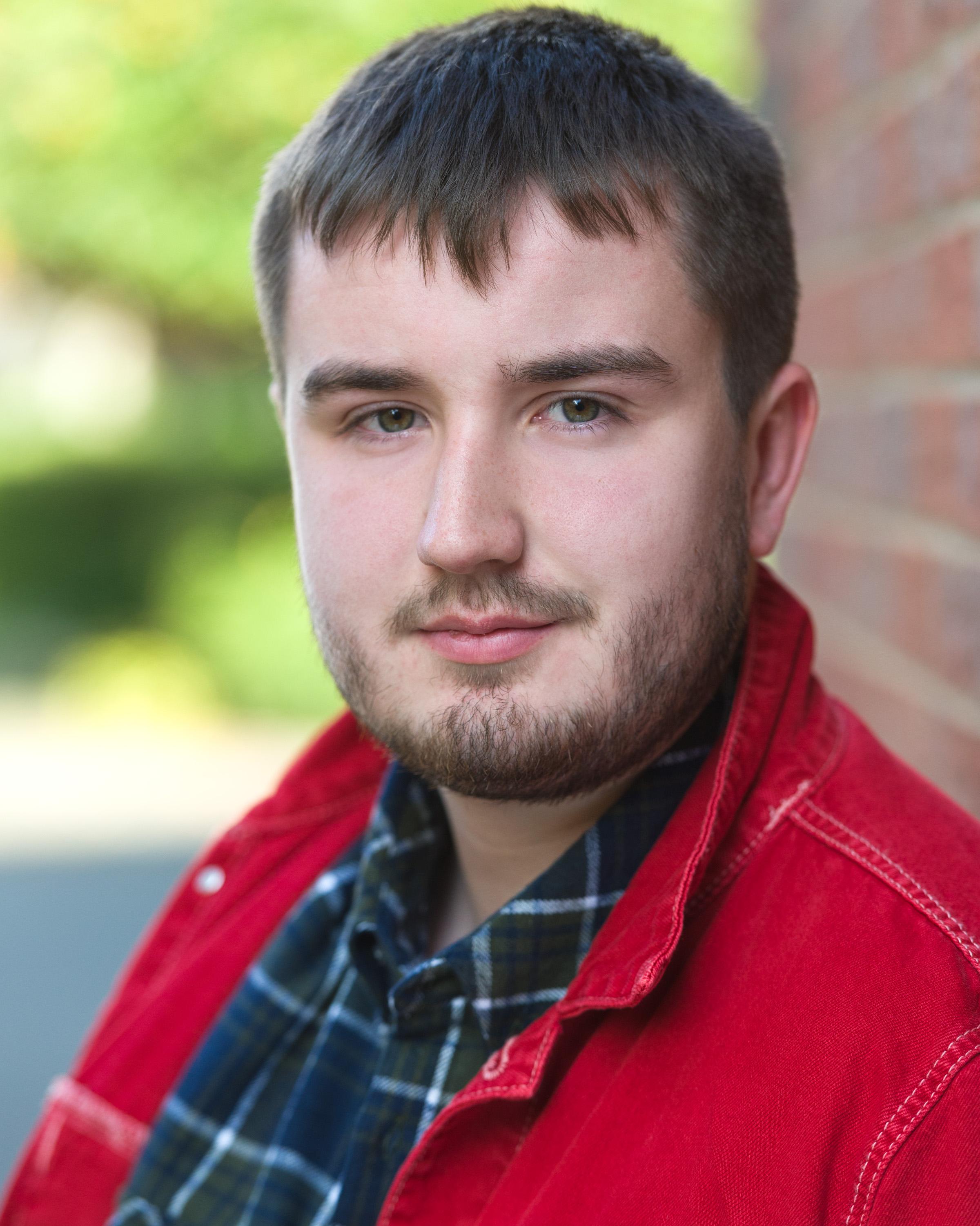 Man with short brown hair, hazel eyes, facial hair wearing a check shirt with a red jacket