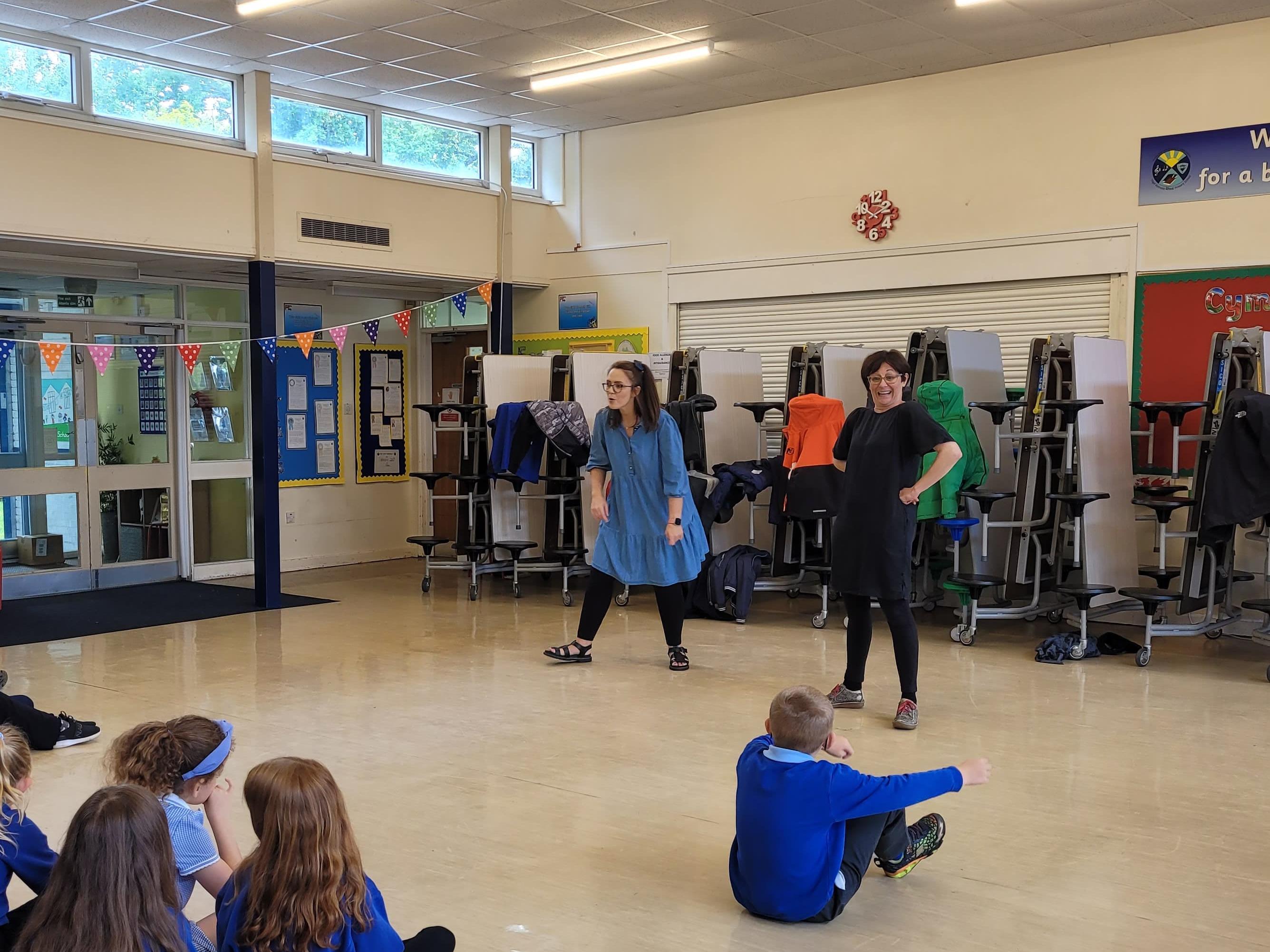 Two women stood in front of children leading a session in a school hall