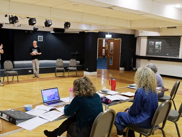 Rehearsal room with two actors and stage managers sat behind a desk
